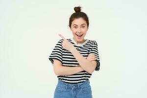Image of excited young woman, smiling, showing amazing price discounts, pointing left, demonstrating something awesome, standing over white background photo