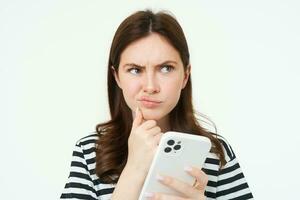 Portrait of girl with complicated face, holds smartphone and looks perplexed, thinks, makes difficult choice or decision, white background photo