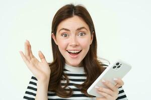 Portrait of happy, surprised young woman, holding mobile phone, showing amazed reaction to great news, standing with smartphone over white background photo