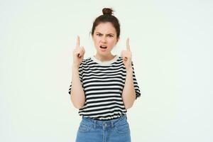 Portrait of angry, frowning woman, arguing and pointing fingers up, stands over white background photo