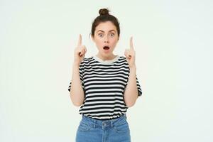 Portrait of surprised girl, opens mouth, says wow, impressed by product on top, points fingers up, stands over white background photo