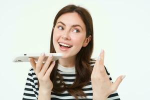 Image of cute brunette woman talking into speakerphone, holding mobile phone near mouth, records her voice, sends a voicemessage, using online translator app, white background photo