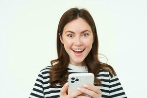 Portrait of excited brunette girl, reacting to amazing promo on smartphone, holding mobile phone and smiling, isolated on white background photo