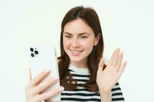 Portrait of woman saying hi, looking at smartphone and waving, video chatting, recording a video blog, connects to online webinar on mobile phone app photo