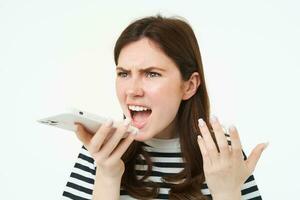 retrato de enojado mujer gritos a teléfono inteligente altavoz, grabación voz mensaje con irritado cara expresión, gritando a teléfono, blanco antecedentes foto