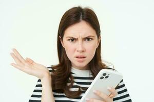 Portrait of confused young woman, looks frustrated, shrugs shoulders, hold mobile phone in one hand, white isolated background photo
