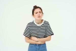 Image of young woman holding hands on her belly, feeling discomfort, menstrual period cramps, needs painkillers, stands over white background photo