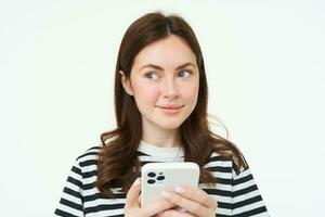 retrato de niña con teléfono, mujer participación teléfono inteligente y pensamiento, sonriente y mirando aparte, decidiendo a comprar algo en línea, blanco antecedentes foto