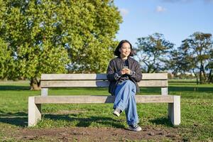Young woman sitting alone on bench in park, using mobile phone, looking at screen, texting, sending messages in app chat photo