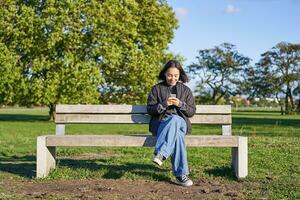 Young woman sitting alone on bench in park, using mobile phone, looking at screen, texting, sending messages in app chat photo