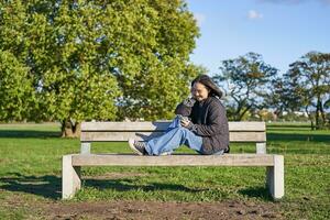 Young woman sitting alone on bench in park, using mobile phone, looking at screen, texting, sending messages in app chat photo