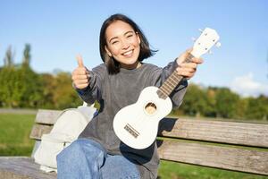 Happy asian girl shows ukulele and thumbs up, demonstrates her new musical instrument, learns how to play in park, sits on bench photo