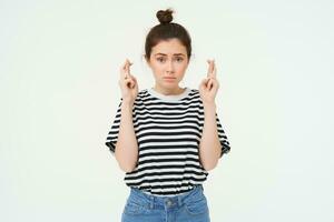 High hopes. Young woman cross fingers, makes wish, anticipates something, stands over white background photo