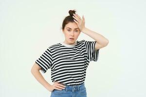 Shocked woman, slaps her forehead, holds hand on head and looks startled, concerned re something bad, terrible situation, standing over white background photo