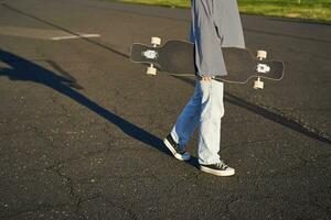 Cropped shot of teen girl body, holding cruiser longboard in hand, walking in sneakers on road in jeans and sweatshirt. Young woman skater with skateboard photo