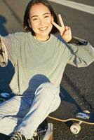 selfie de asiático niña sentado en patineta, tomando foto en teléfono inteligente, sonriente y demostración paz signo v