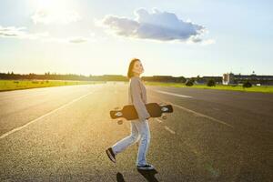 Asian girl with skateboard standing on road during sunset. Skater posing with her long board, cruiser deck during training photo