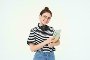 Education concept. Smiling brunette girl, student in casual clothes, holds her books, study material, wears headphones over neck, looks confident and happy, isolated over white background photo