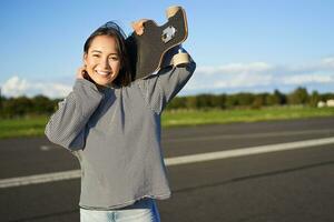 retrato de hermosa joven patinador chica, en pie con longboard y sonriente a cámara. asiático mujer con patineta en pie en la carretera foto