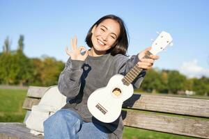 linda sonriente niña muestra Okay firmar y su nuevo ukelele, se sienta en banco en parque, recomienda musical instrumento foto