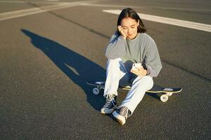 retrato de asiático mujer sentado en patineta, Patinaje en su crucero longboard, utilizando teléfono inteligente aplicación foto