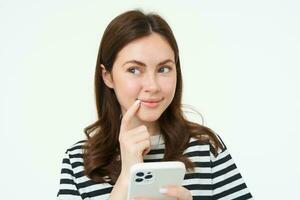 Portrait of smiling woman thinks to order something online on smartphone app, holding mobile phone with thoughtful face photo