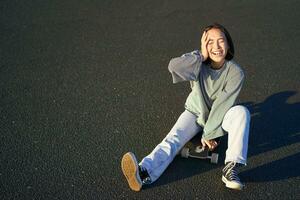 contento hermosa coreano adolescente niña se sienta en su patineta, crucero en longboard, vistiendo casual ropa foto