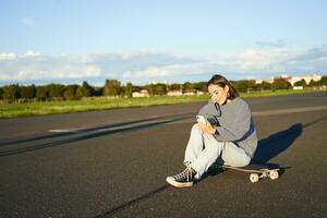 Portrait of young korean girl sitting on her skateboard on road, looking at smartphone, chatting on mobile app photo