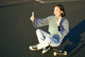 Positive teen asian girl takes selfie, video chats on mobile phone app, sits on skateboard and records vlog photo