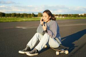 retrato de asiático niña se sienta en su longboard, Patinaje en patineta y hablando en móvil teléfono, teniendo conflicto durante teléfono conversación, discutiendo con preocupado cara foto