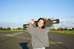 Leisure and people. Happy asian woman standing with longboard, cruising on an empty road in countryside. Skater girl holds her skateboard and smiles at camera photo