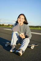 Vertical shot of asian woman sitting on skateboard on road, holding smartphone app. Skater girl skates on longboard, using mobile phone photo