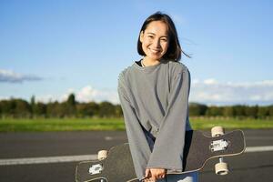 retrato de hermosa asiático niña Patinaje en longboard, cruzando con patineta en vacío camino, disfrutando tiempo libre en Fresco aire foto