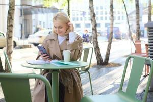 Street style portrait of young smiling woman sitting on street outdoors, holding mobile phone, using smartphone, drinking coffee in local cafe photo