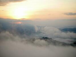 The mist flows through the mountain forest, Sun shining into tropical forest, Mist drifts through mountain ridges in the morning, slow floating fog blowing cover on the top of mountain photo