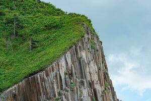 fragment of a cliff made of columnar basalt with lush grass on top against the sky photo