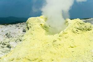 cone of sulfur deposits around a fumarole in a solfataric field illuminated by the sun against a stormy landscape in the distance photo