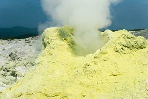 cone of sulfur deposits around a fumarole in a solfataric field illuminated by the sun against a stormy landscape in the distance photo