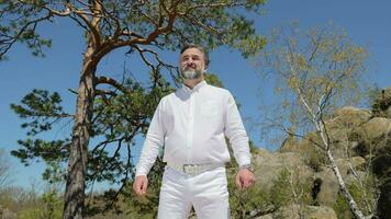 Man in White Outfit Outdoor Portrait. Man dressed in white stands on top of a mountain against the background of pine trees on a sunny day. Portrait of the artist. A man with a beard. video