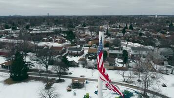 ein Drohne fliegt um das uns Flagge im Winter im Illinois. das USA Flagge fliegt im das Center von das Dorf im wolkig Wetter im Winter. video