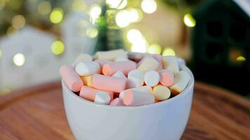 A cup of hot chocolate with marshmallows on a wooden table surrounded by Christmas lights. A close-up of a white cup with a hot drink sprinkled with marshmallows on top. video