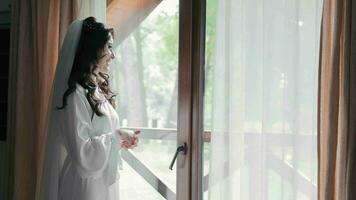 Wedding morning of the bride. A happy bride is standing by the window in a white robe, hoping to see the groom through the window. The bride looks out the window and fixes her hair. video