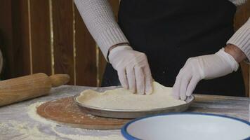 A man in an apron is spreading tomato paste on a pizza dough base on a table sprinkled with flour. On the table in front of him are bowls of pizza ingredients. He pinches the edges of the dough with video