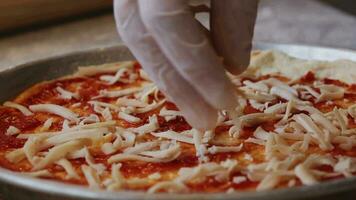 A man in an apron is pouring grated cheese onto a pizza dough base on a table sprinkled with flour. On the table in front of him are bowls of pizza ingredients. He pinches the edges of the dough with video