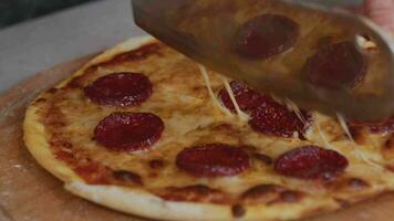 ein Mann im ein Schürze zieht Pizza aus von ein holzbefeuert Ofen. ein Koch zieht aus ein gekocht Pizza. Verbrennung Essen im ein holzbefeuert Ofen. groß Messer schneidet Pizza mit Käse. Kochen Essen Über ein öffnen Feuer. natürlich video