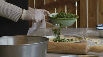 A man in an apron is spreading tomato paste on a pizza dough base on a table sprinkled with flour. On the table in front of him are bowls of pizza ingredients. He pinches the edges of the dough with video