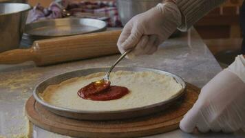 A man in an apron is spreading tomato paste on a pizza dough base on a table sprinkled with flour. On the table in front of him are bowls of pizza ingredients. He pinches the edges of the dough with video