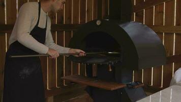 A man in an apron is putting raw pizza into a wood-fired pizza oven. Cooking food over an open fire. Natural food. Neapolitan pizza in the oven. Crumbling cheese on the surface of the pizza. video