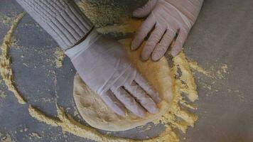 A man in an apron is spreading tomato paste on a pizza dough base on a table sprinkled with flour. On the table in front of him are bowls of pizza ingredients. He pinches the edges of the dough with video