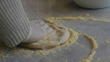 A man in an apron is spreading tomato paste on a pizza dough base on a table sprinkled with flour. On the table in front of him are bowls of pizza ingredients. He pinches the edges of the dough with video